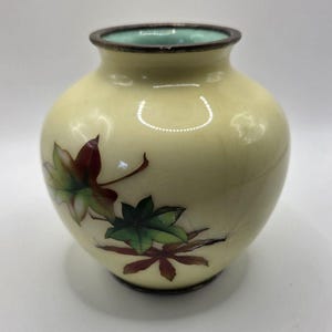 May include: A small, round, cream-colored vase with a smooth, glossy finish. The exterior features a painted design of maple leaves in shades of deep red, brown, and green. The rim and base are trimmed with a dark, metallic-toned band.