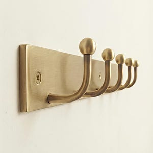 Puede incluir: Un perchero de pared con una placa rectangular de latón cepillado y cinco ganchos curvos. Cada gancho termina en un pomo esférico. La placa está asegurada con tornillos visibles en ambos extremos, mostrando un acabado metálico y diseño lineal.