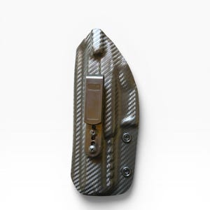May include: A carbon fiber-patterned holster with a metal clip and screws. The holster is dark gray and black, with a textured surface. The clip is a light brown color. The holster is designed to be worn on the body.