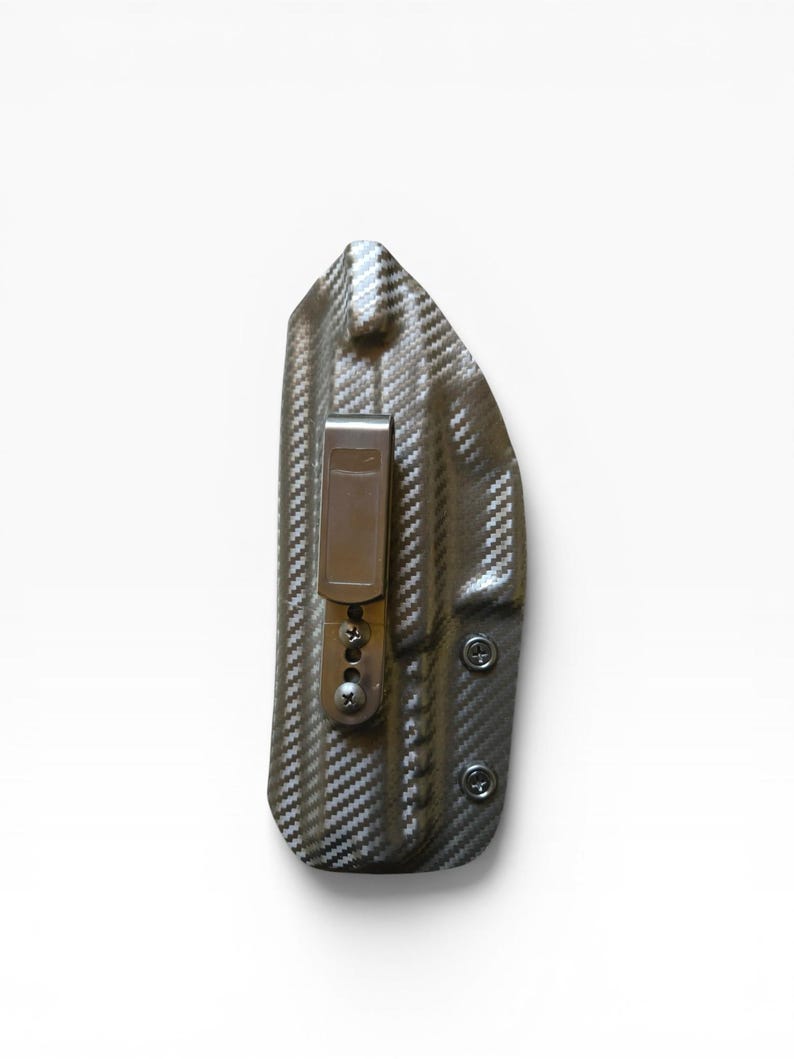 May include: A carbon fiber-patterned holster with a metal clip and screws. The holster is dark gray and black, with a textured surface. The clip is a light brown color. The holster is designed to be worn on the body.