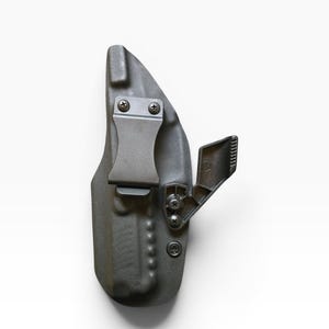 May include: A dark gray, molded polymer holster with a metal belt clip and adjustable retention device. The holster is designed to be worn on the body and has several screws for adjustment and assembly. The overall design is compact and functional.