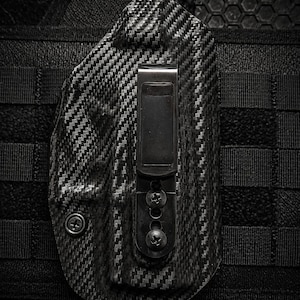 May include: A black carbon fiber holster with a metal clip and screws. The holster is attached to a black tactical vest with a textured pattern. The holster has a woven carbon fiber appearance.