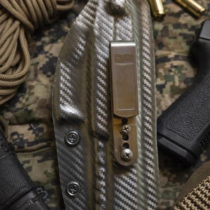 May include: A gray and black patterned holster with a metal clip and screws. The holster is designed to hold a firearm. The background includes ammunition, a pistol, and a camouflage pattern.