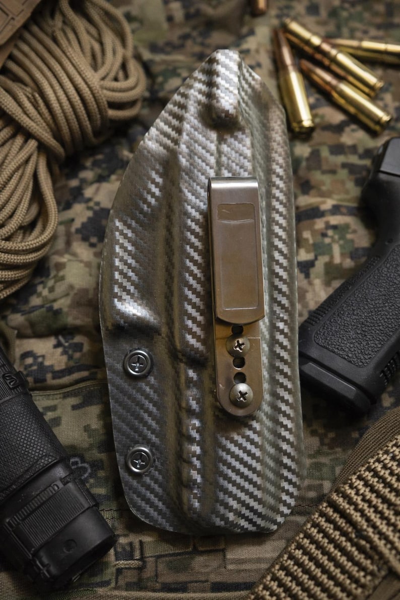 May include: A gray and black patterned holster with a metal clip and screws. The holster is designed to hold a firearm. The background includes ammunition, a pistol, and a camouflage pattern.