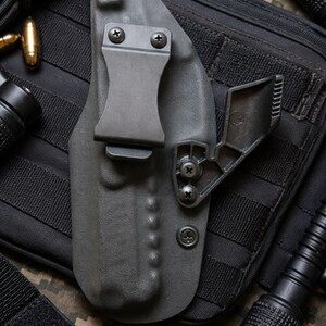May include: A gray, molded polymer holster with a metal belt clip and adjustable retention device. The holster is designed to securely hold a firearm. The background includes a black tactical pouch, flashlight, and ammunition.