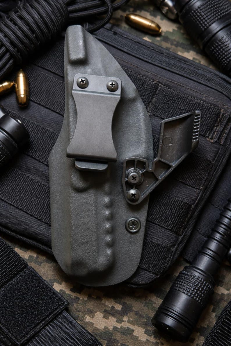 May include: A gray, molded polymer holster with a metal belt clip and adjustable retention device. The holster is designed to securely hold a firearm. The background includes a black tactical pouch, flashlight, and ammunition.