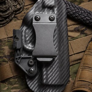 May include: A black carbon fiber holster with a textured pattern, featuring a metal clip and screws. The holster is set against a camouflage background, with a black rope and other tactical gear visible.