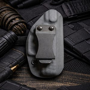 May include: A gray polymer holster with a metal clip and screws. The holster is designed to hold a firearm. The background includes black tactical gear and ammunition.