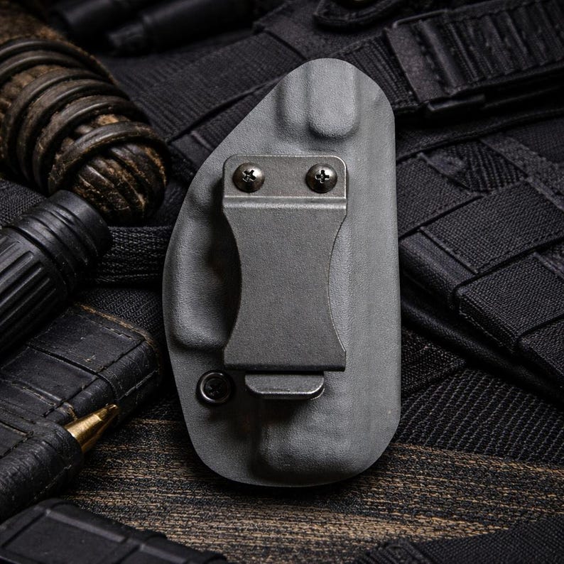 May include: A gray polymer holster with a metal clip and screws. The holster is designed to hold a firearm. The background includes black tactical gear and ammunition.