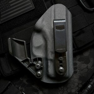May include: A black molded holster featuring a smooth matte finish, a sturdy metal belt clip, and an adjustable claw attachment secured with two screws. The holster is constructed from a rigid polymer material with a contoured shape for retention.