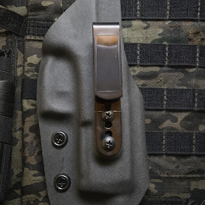 May include: A gray, molded polymer holster with a metal clip and screws. The holster is attached to a camouflage fabric background. The clip is a dark bronze color. The holster has two black, circular fasteners.