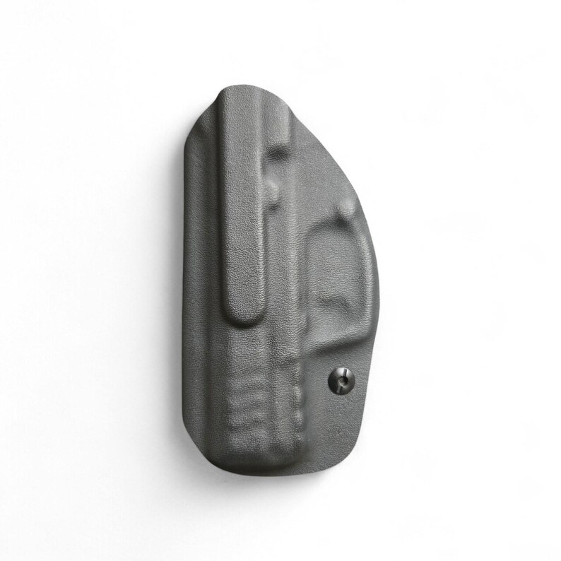 May include: A gray, molded polymer holster with a belt clip and a single screw. The holster is designed to be worn on the body. The surface has a textured finish.