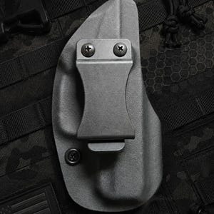 May include: A gray, molded polymer holster with a metal belt clip, secured by two screws. The holster is designed for concealed carry and is shown on a tactical background with an American flag patch.