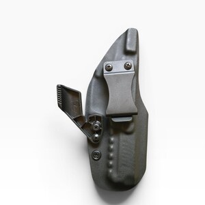 May include: A dark gray, molded polymer holster with a metal belt clip and mounting hardware. The holster features a curved design and is secured with screws. The clip is designed to attach to a belt for concealed carry.