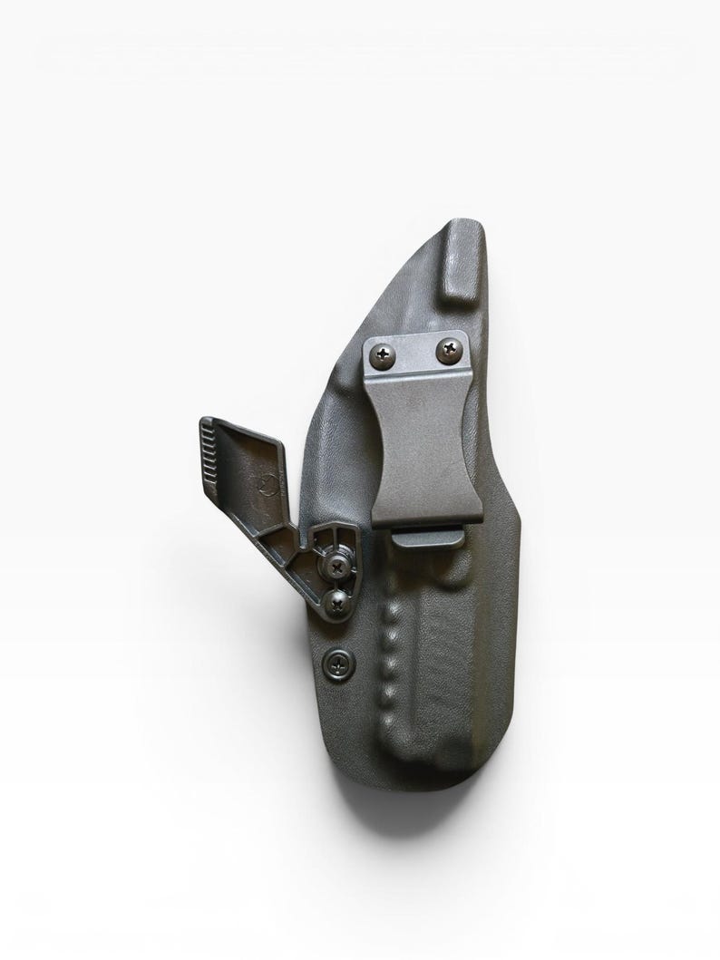 May include: A dark gray, molded polymer holster with a metal belt clip and mounting hardware. The holster features a curved design and is secured with screws. The clip is designed to attach to a belt for concealed carry.