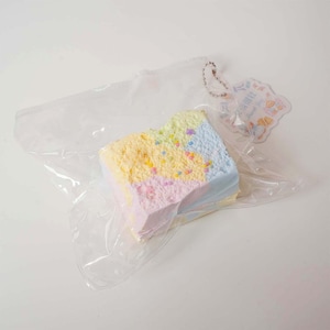May include: A pastel-colored, square-shaped item resembling a slice of cake, packaged in a clear plastic bag. The cake has layers of yellow, pink, and blue with colorful sprinkles. A small tag with text hangs from a chain.