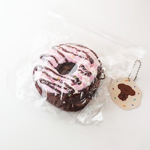 May include: A chocolate-frosted donut with pink icing and chocolate drizzle, packaged in clear plastic. A small tag with a cartoon dog illustration is attached. The donut is a whimsical treat.