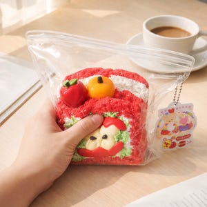 May include: A red and white cake roll with fruit-shaped decorations, held in a clear plastic bag. The cake has a decorative apple design. A small tag with cartoon characters hangs from the bag.