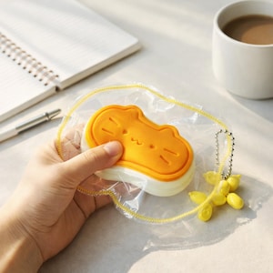 May include: A yellow and white cat-shaped object is held in a clear plastic pouch with a yellow zipper. The cat has a smiling face. A yellow charm with a chain is attached to the pouch.