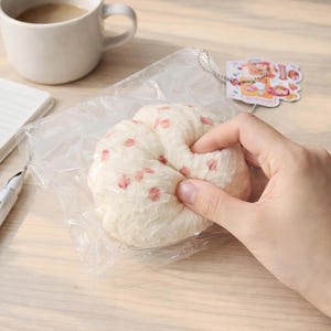 May include: A soft, white, round plush toy featuring a delicate pink cherry blossom petal pattern. The plush is encased in a clear plastic bag and includes a small, rectangular paper tag attached by a silver-colored metal ball chain.