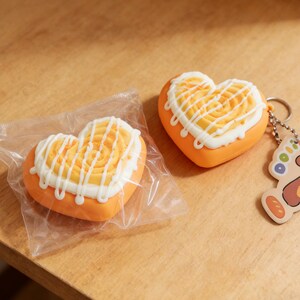 May include: Two heart-shaped objects with orange bases and yellow tops, decorated with white icing. One is in a clear plastic bag, the other is a keychain. The keychain has a silver ring and a small charm.