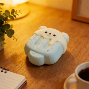May include: A light blue, bear-shaped night light resembling an ice cream bar. The top is white with a dripping effect, and the bear has small brown eyes and a red mouth. The light is on a wooden surface.