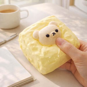 May include: A yellow, cube-shaped item with a white bear face in the center. The bear has black eyes and a black cross for a nose. The item is held by a hand, and a white coffee cup and notebook are in the background.