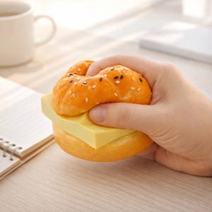 May include: A novelty stress ball shaped like a hamburger. The bun is orange with sesame seeds, and the cheese is yellow. The stress ball is held in a hand, suggesting it's a fun desk accessory or a playful item for stress relief.