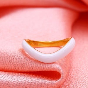 May include: A gold-toned ring featuring a smooth, white enamel inlay along the outer band. The ring has a distinct V-shaped or chevron contour, creating a curved silhouette that sits prominently against the surrounding fabric texture.