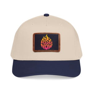 Puede incluir: Gorra de béisbol con corona color crema y visera azul marino. Un parche rectangular de cuero muestra las palabras "RISE NOW" en naranja y rosa, con un diseño de llama.