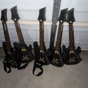 Könnte beinhalten: Eine Sammlung von fünf schwarzen Spielzeug-Gitarren mit braunen Hälsen und schwarzen Gurten. Die Gitarren haben eine markante Form mit einer spitzen Kopfplatte und goldenen Akzenten. Die Gitarren sind für ein Videospiel konzipiert.