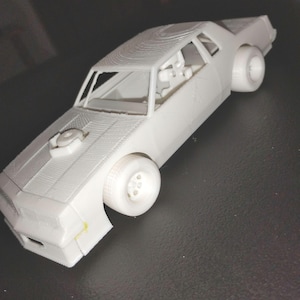 May include: A white 3D-printed model car with a detailed exterior, including wheels, windows, and a hood scoop. The car is set against a dark background, showcasing its design and form.