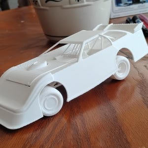 Può includere: Un modellino di auto da corsa stampato in 3D di colore bianco, dotato di un prominente alettone posteriore, una gabbia di sicurezza dettagliata visibile attraverso i finestrini aperti e quattro ruote modellate.