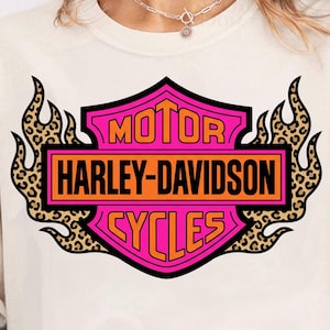 May include: A cream-colored t-shirt featuring a graphic of the Harley-Davidson Motor Cycles logo in bright pink and orange. The logo is framed by stylized flames with a brown and black leopard print pattern on a light-colored background.