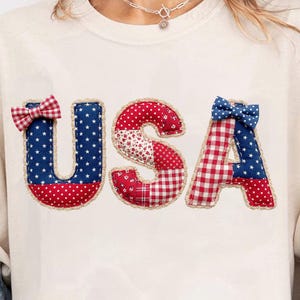 Könnte beinhalten: Cremefarbenes T-Shirt mit dem Wort "USA" aus rot, weiß und blauem Stoff. Die Buchstaben sind mit Sternen, Punkten und Vichy-Mustern verziert, und jeder Buchstabe hat eine kleine Schleife. Das Shirt ist ein lässiger Stil.