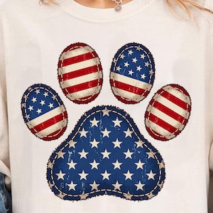 Puede incluir: Camiseta color crema con un diseño de huella de pata patriótica. La huella de la pata está compuesta por estrellas azules y blancas y rayas rojas y blancas, que se asemejan a la bandera estadounidense. El diseño está cosido con hilo azul oscuro.
