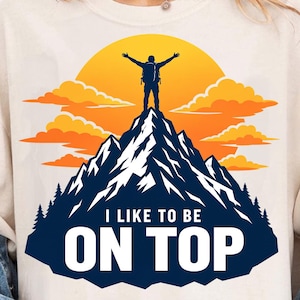 May include: A cream-colored t-shirt featuring a graphic of a hiker standing on a mountain peak with arms raised. The design includes a large yellow sun, orange clouds, dark blue mountains, and the text I LIKE TO BE ON TOP in white letters.