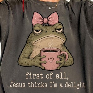 May include: A charcoal gray t-shirt featuring a graphic of a green toad wearing a pink polka-dot bow, holding a pink mug with a heart icon. Below the toad, the text reads: first of all, Jesus thinks I'm a delight in a simple white font.