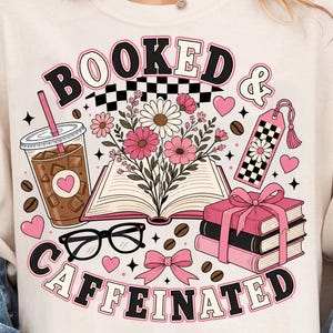 Puede incluir: Una camiseta color crema con un estampado gráfico que incluye el texto BOOKED & CAFFEINATED. El diseño muestra un libro abierto con flores, un vaso de café con pajita, una pila de libros con un lazo rosa, gafas negras y un marcapáginas a cuadros.