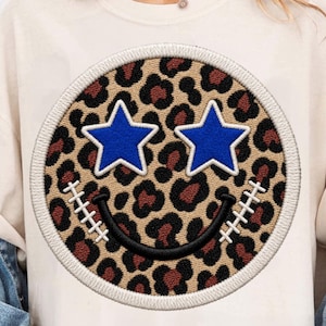 May include: A cream-colored t-shirt featuring a large circular patch with a brown and black leopard print pattern. The patch displays two bright blue star-shaped eyes and a black curved smile with white stitched accents on each side.