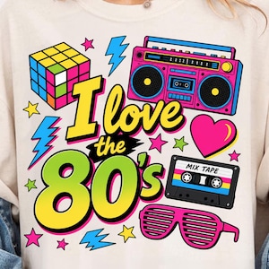 Pode incluir: Uma t-shirt de cor creme com uma impressão gráfica colorida com o texto "I love the 80's". O design inclui um cubo de Rubik, um boombox cor-de-rosa, uma cassete com a etiqueta "MIX TAPE", óculos de persiana, um coração, estrelas e raios.