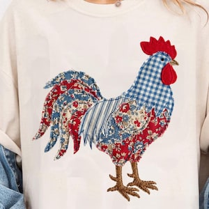 Peut inclure: T-shirt crème avec un appliqué de coq. Le coq a une crête et un fanon rouges, un corps à carreaux bleu et blanc, et une queue avec des motifs floraux et rayés rouges, blancs et bleus. Les pattes du coq sont marron.