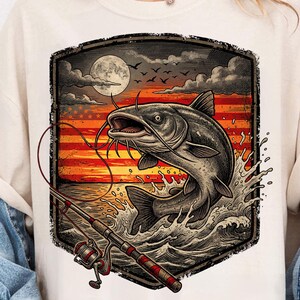 May include: Cream-colored t-shirt featuring a graphic of a catfish leaping from water, with a fishing rod in the foreground. The background shows an American flag design with a sunset, moon, and birds. The design is framed in a dark border.