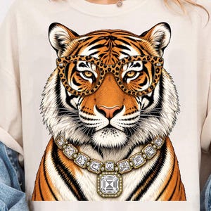 Könnte beinhalten: Ein cremefarbenes T-Shirt mit einem Grafikdruck eines Tigers, der eine Brille mit Leopardenmuster und eine große, facettierte Kristallkette trägt. Der Tiger hat orangefarbene und schwarze Streifen mit weißem Fell.