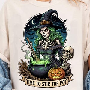 May include: A cream-colored t-shirt featuring a graphic of a green-haired skeleton witch in a black hat stirring a cauldron. The design includes a skull, a pumpkin, bats, a crescent moon, and the text "TIME TO STIR THE POT" on a banner.