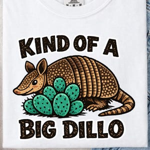 May include: White t-shirt with a graphic of an armadillo and cactus, with the text "KIND OF A BIG DILLO" in black. The armadillo is brown and tan, and the cactus is green.