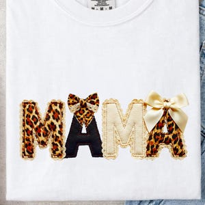 Puede incluir: Camiseta blanca con la palabra "MAMA" aplicada en la parte delantera. Las letras son una mezcla de estampado de leopardo, negro y crema, con lazos decorativos y ribete de encaje. La camiseta es de material suave.