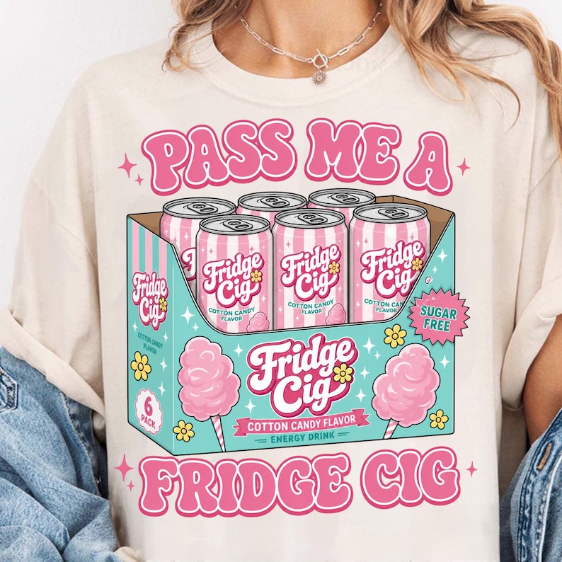 Op de afbeelding: Een cr&egrave;mekleurig t-shirt met een afbeelding van een sixpack roze Fridge Cig energiedrankblikjes in een blauwgroene doos. Het ontwerp bevat de tekst PASS ME A FRIDGE CIG, COTTON CANDY FLAVOR en SUGAR FREE, versierd met roze suikerspinillustraties.