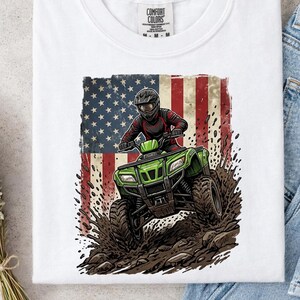 May include: White t-shirt with a graphic of a person riding a green ATV, set against an American flag. The ATV is shown kicking up mud. The shirt has a Comfort Colors tag at the top.