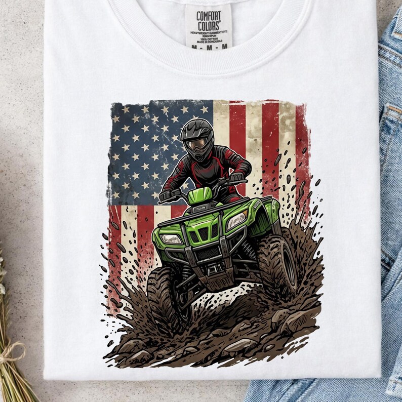 May include: White t-shirt with a graphic of a person riding a green ATV, set against an American flag. The ATV is shown kicking up mud. The shirt has a Comfort Colors tag at the top.